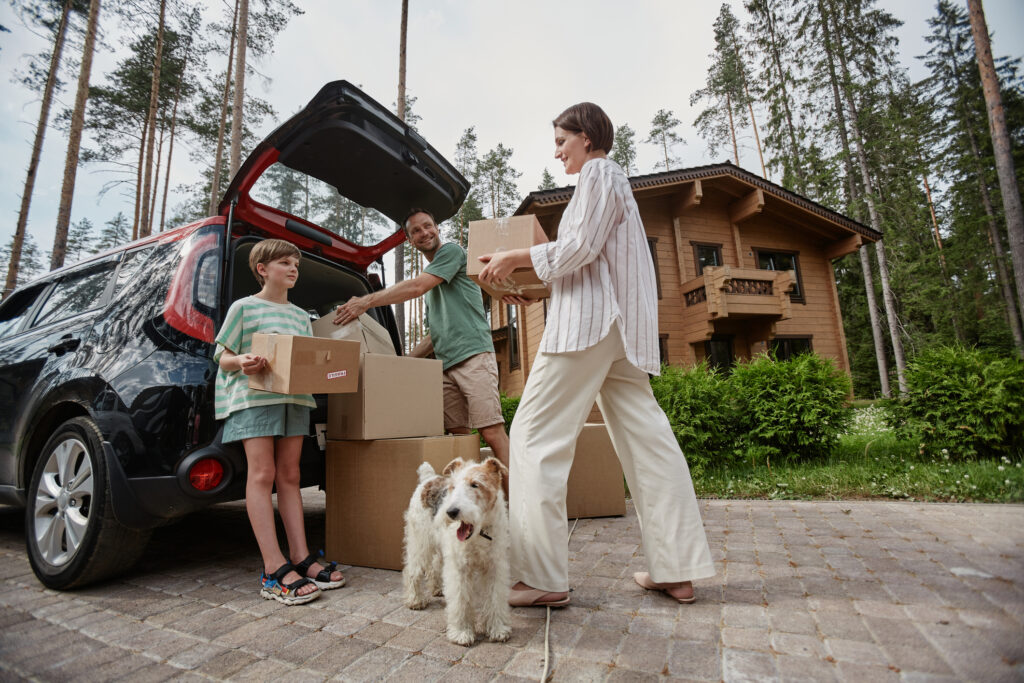 A family PCSing to a new state, unloading boxes from their car in front of a wooden house, with their child holding a box