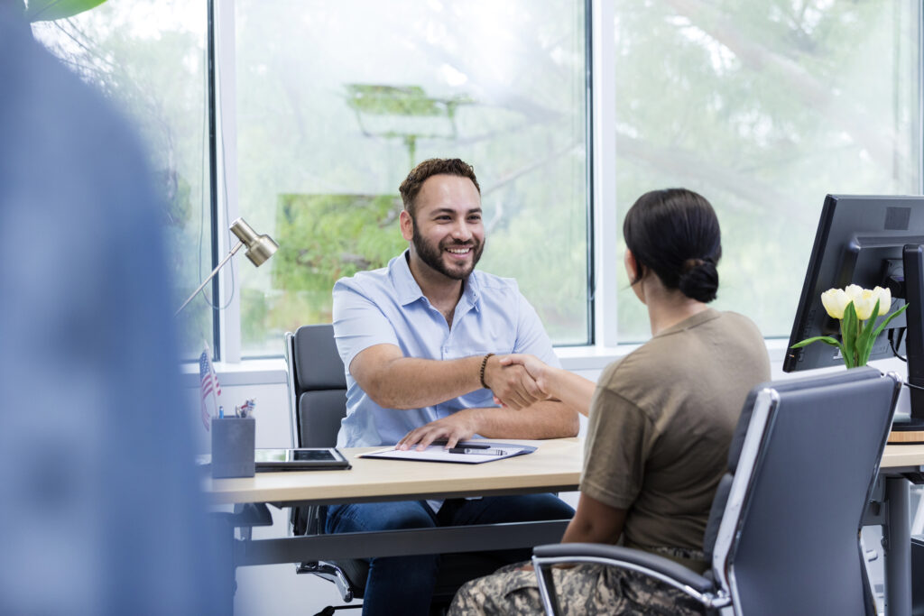 Military service member meeting with a smiling professional at a desk, shaking hands in a bright office environment.