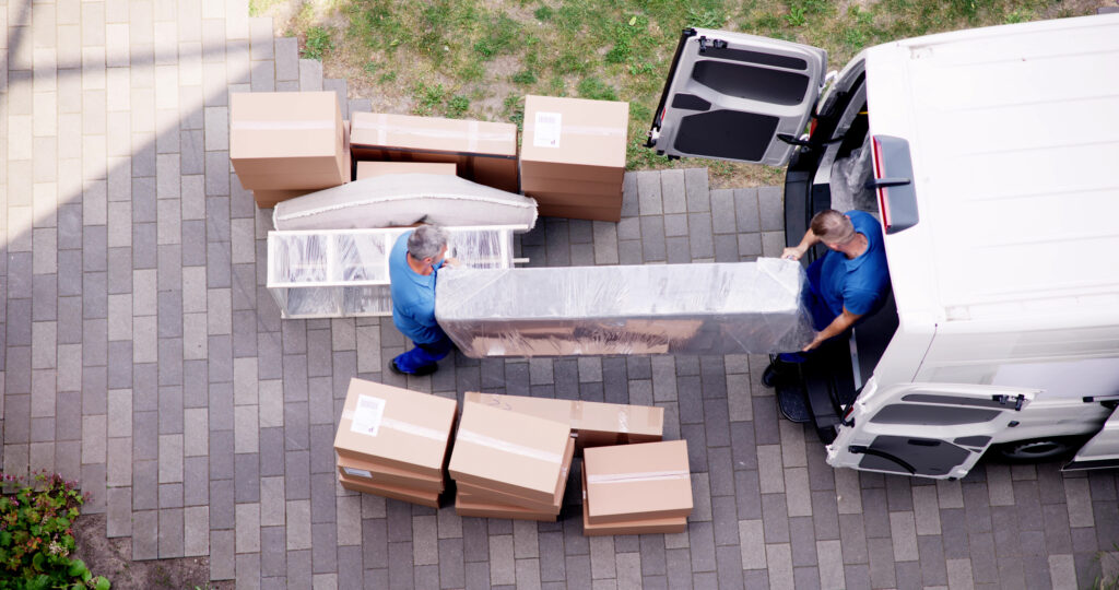 Two movers unloading wrapped furniture and cardboard boxes from a white van onto a paved driveway.