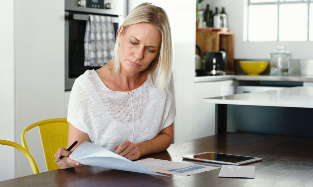 A woman sits at a kitchen table, reviewing and filling out military move claim documents with a pen, surrounded by paperwork and a tablet.